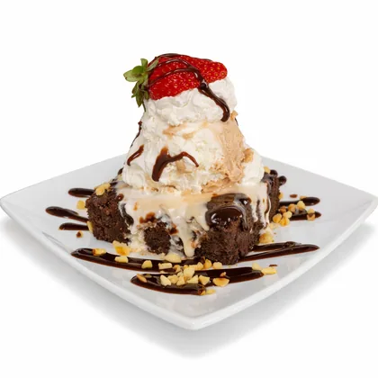 Brownie con bolas de helado, crema, fresas, salsa de chocolate y maní sobre un plato blanco.