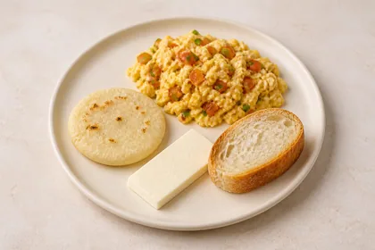 Desayuno con huevo perico, arepa, pan y queso blanco sobre un plato crema.