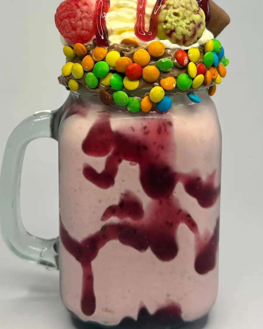 Malteada rosada con crema, galletas, dulces de colores y salsa roja en una jarra de vidrio.
